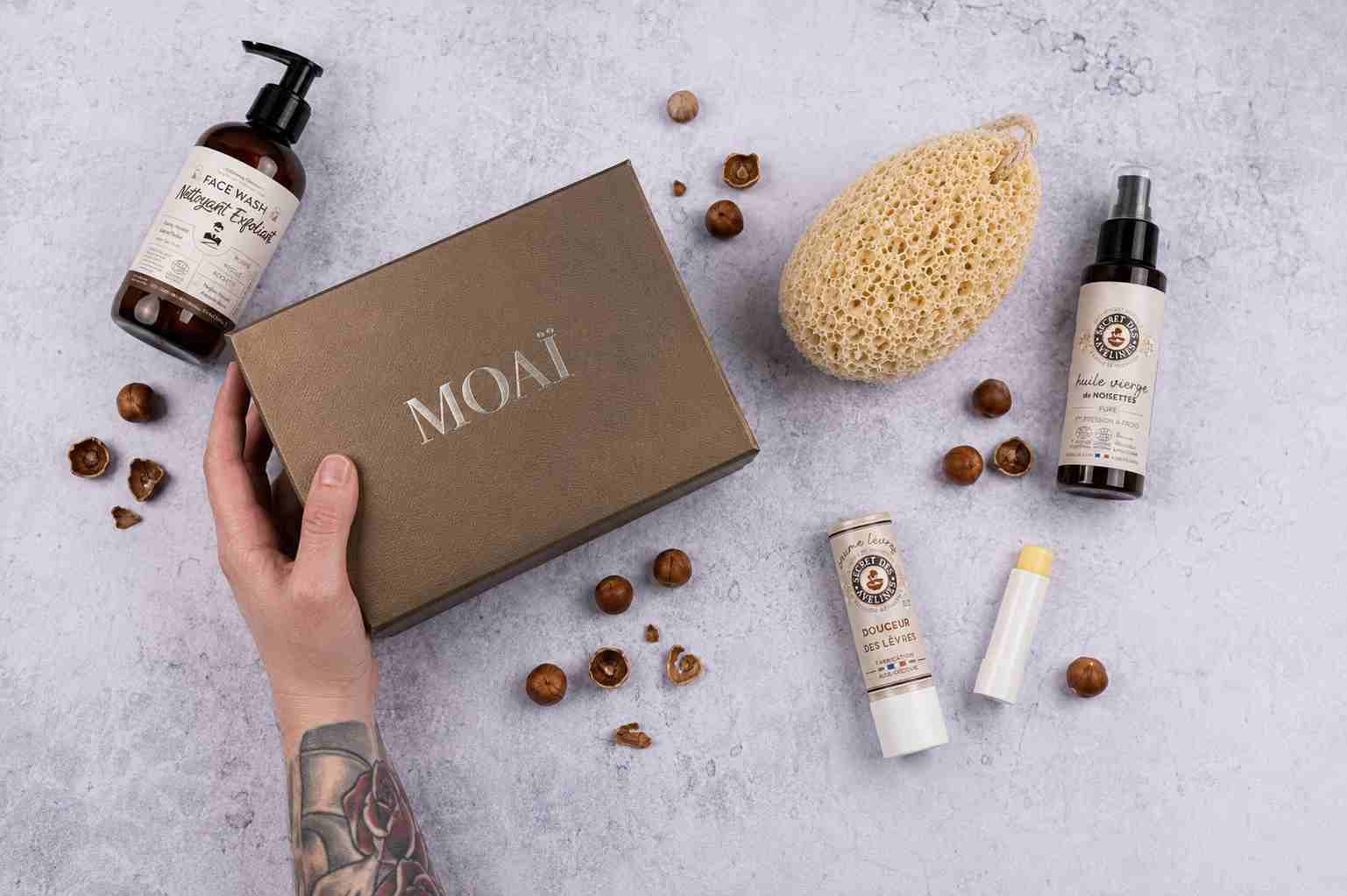 Coffret de soins homme MOAÏ avec nettoyant visage au charbon, huile nourrissante à la noisette, baume à lèvres et éponge naturelle. Présentation à plat avec coffret noir et noisettes.