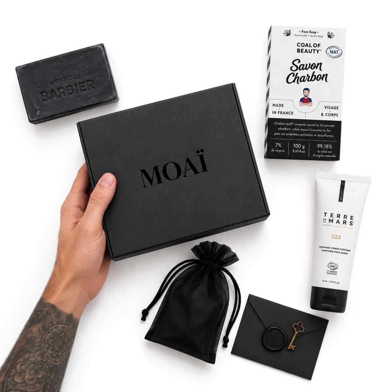 Main tatouée tenant une box noire de la marque MOAÏ entourée d'un gommage Terre de Mars, d'un savon au charbon actif et d'un pochon noir.