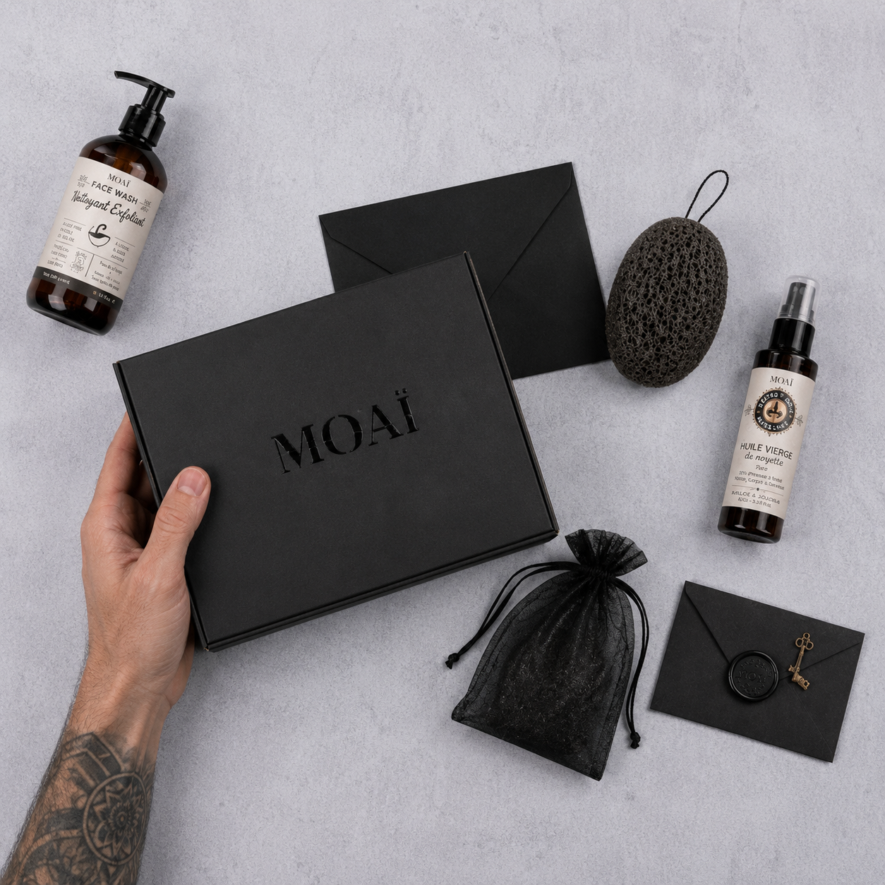 Vue de dessus à plat du coffret cadeau noir MOAÏ tenu en main, entouré du nettoyant nettoyant exfoliant, de l'huile de noisette, de l'éponge konjac et de la pochette d'accessoires.