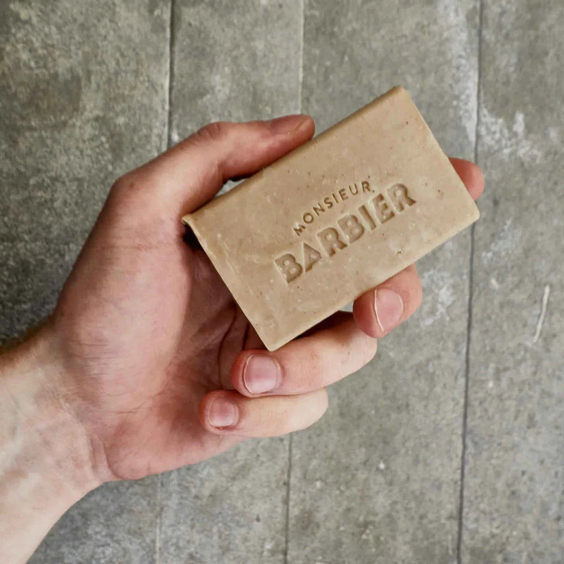 Main d'homme tenant un pain de savon solide gravé au nom de la marque Monsieur Barbier sur un fond texturé gris.
